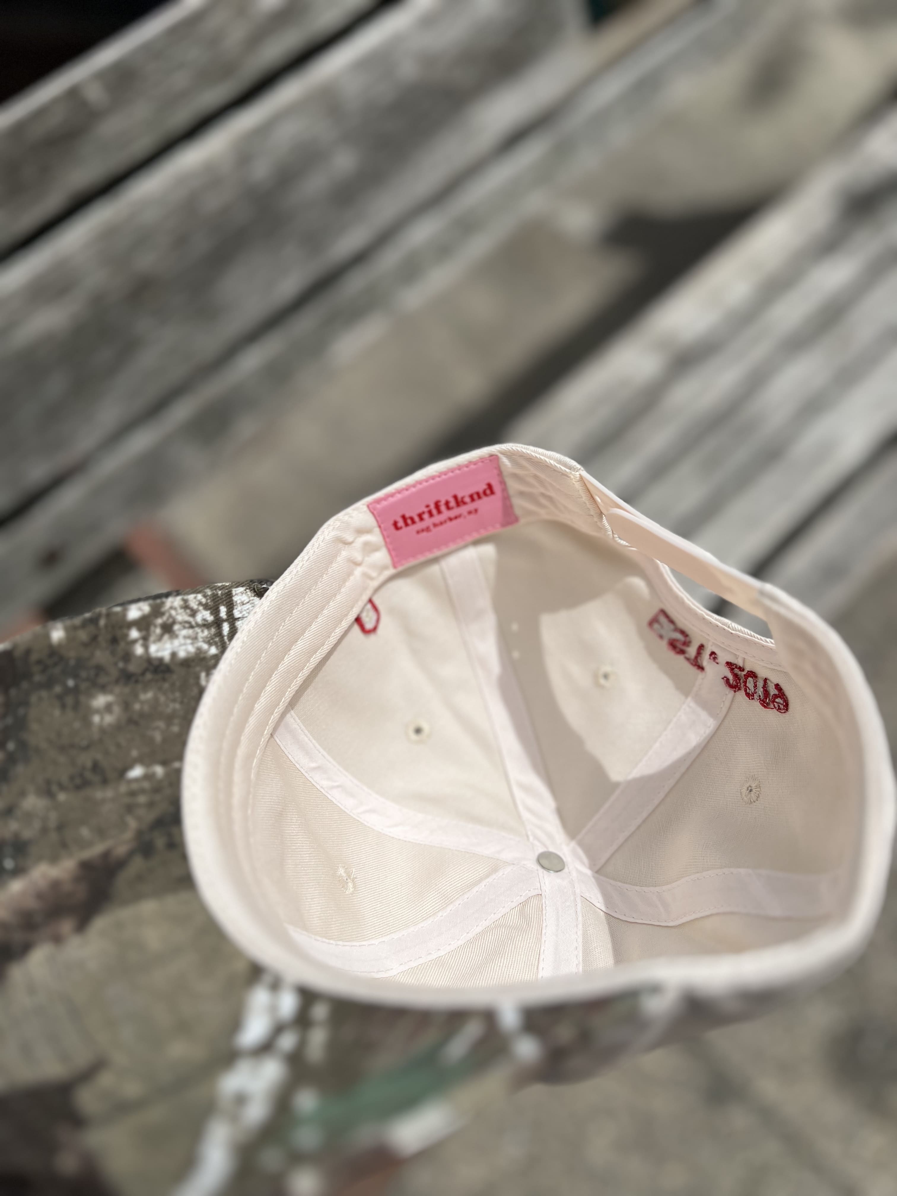 White baseball cap with pink thriftkind label tag visible on interior lining, displayed upside down on wooden surface