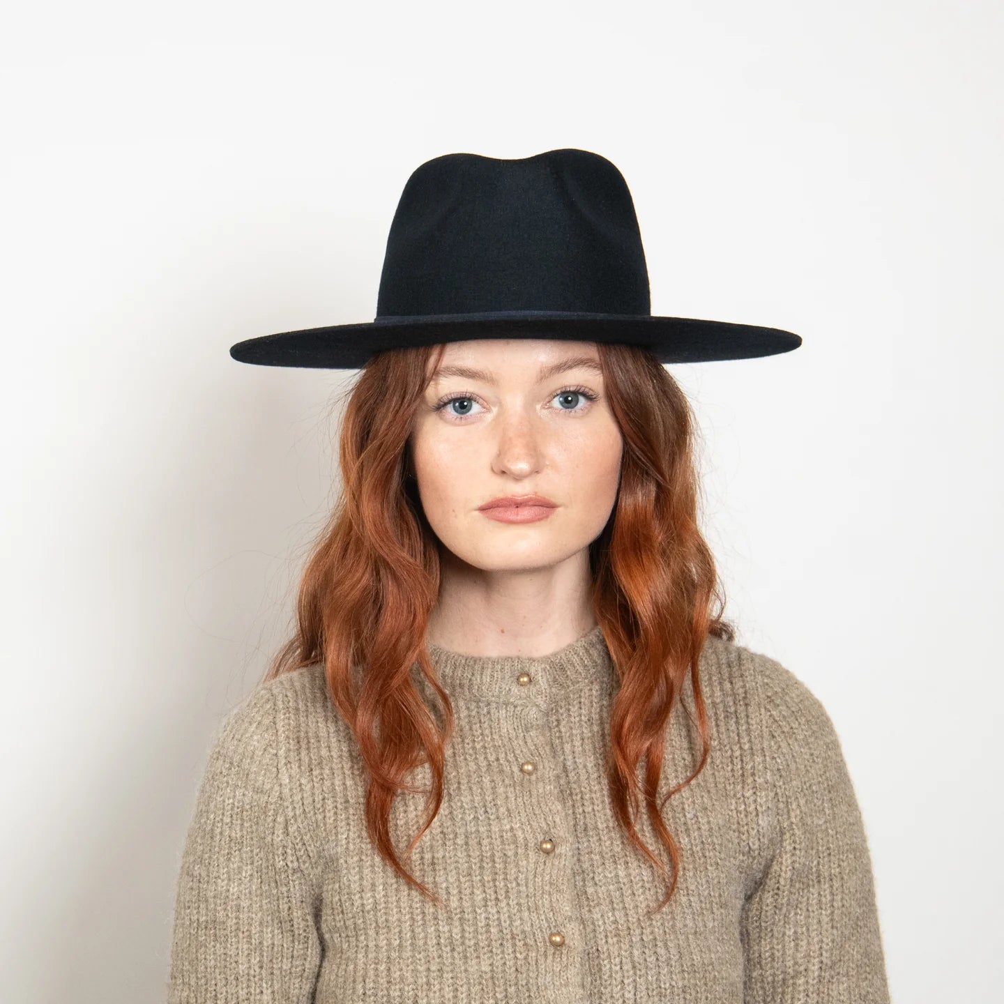 Black wide-brim fedora hat worn by woman with red hair and beige sweater against white background