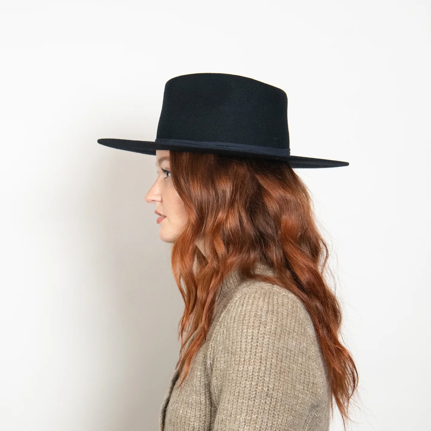 Black wide-brim felt fedora hat with flat crown, modeled on woman with long auburn hair wearing beige sweater, side view