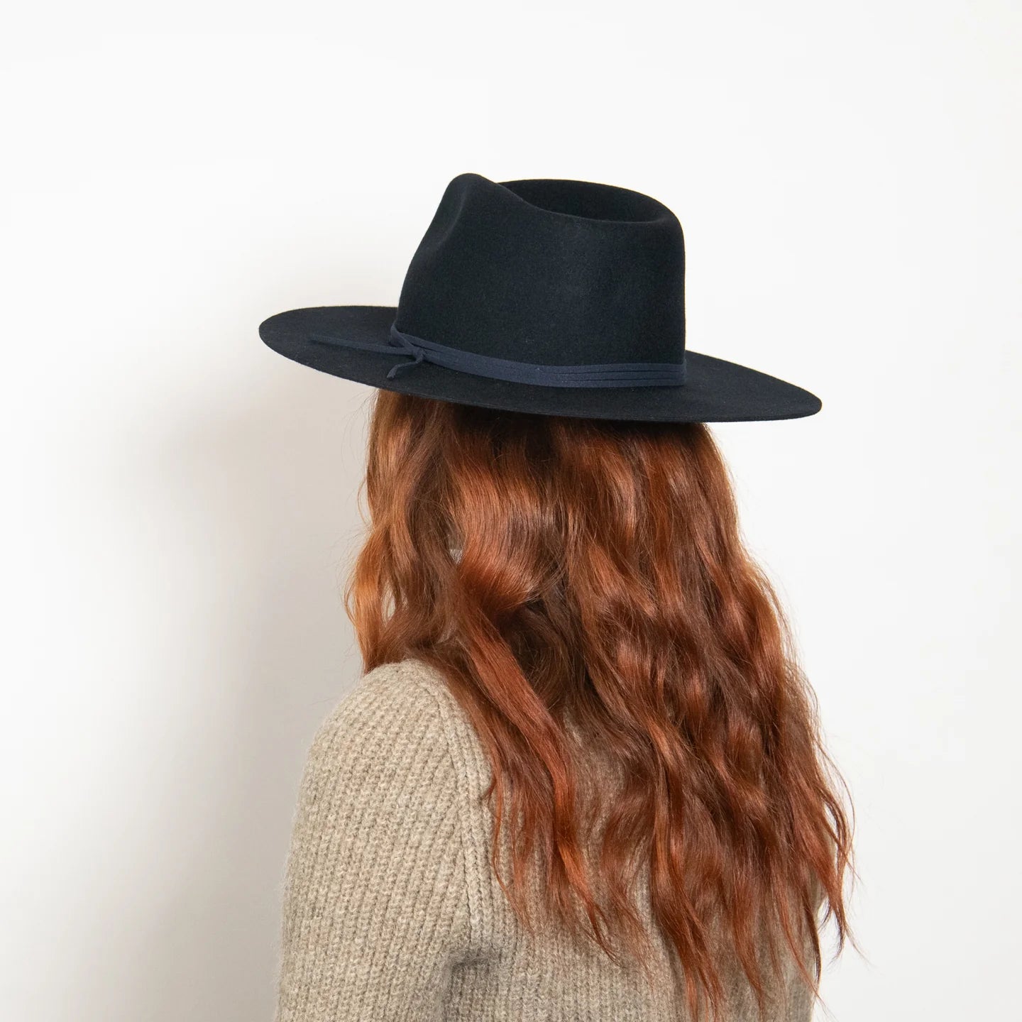 Black felt wide-brim fedora hat with navy ribbon band, shown from back on person with long red hair wearing beige sweater