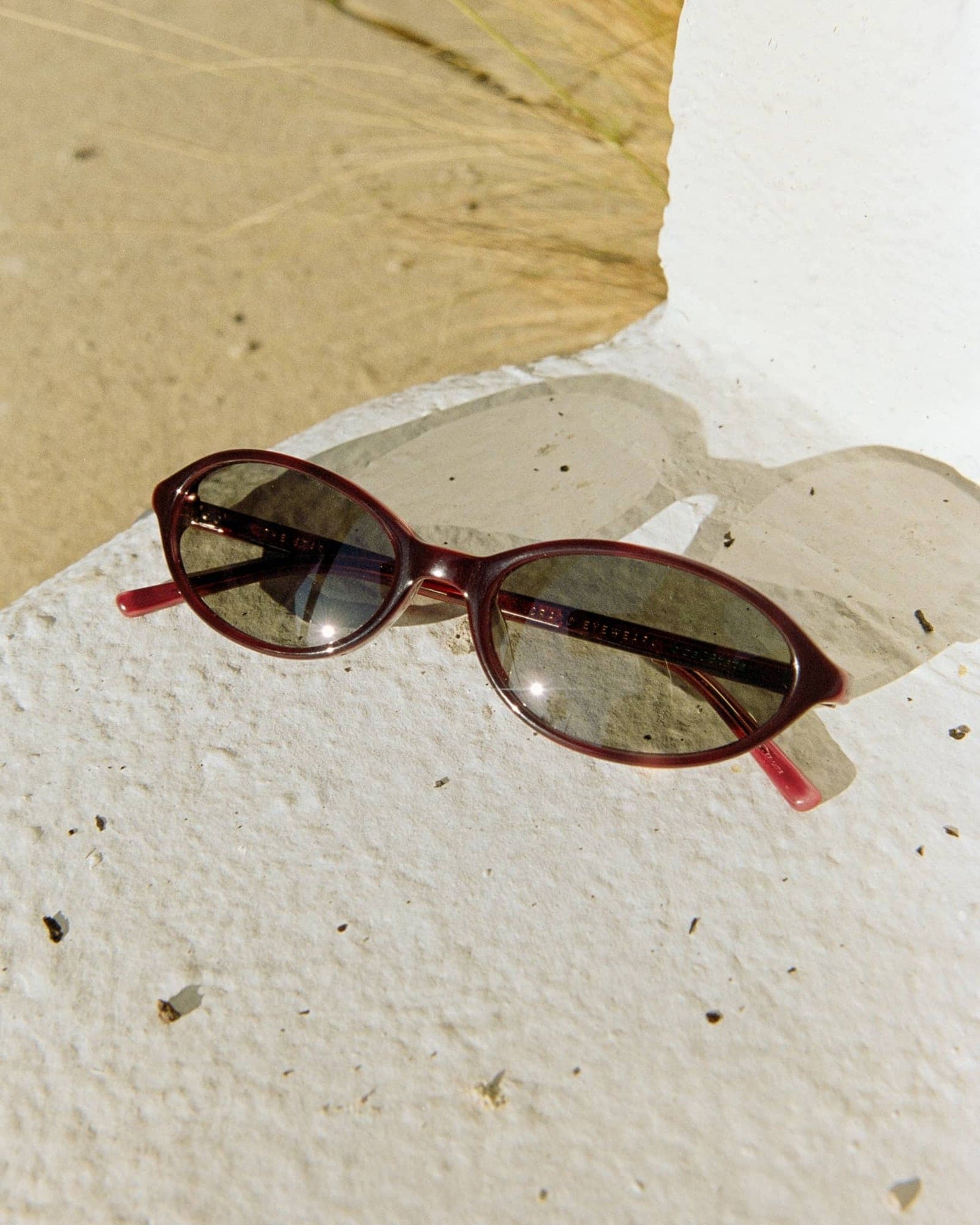 Burgundy red oval sunglasses with gradient lenses on textured white concrete surface with natural lighting