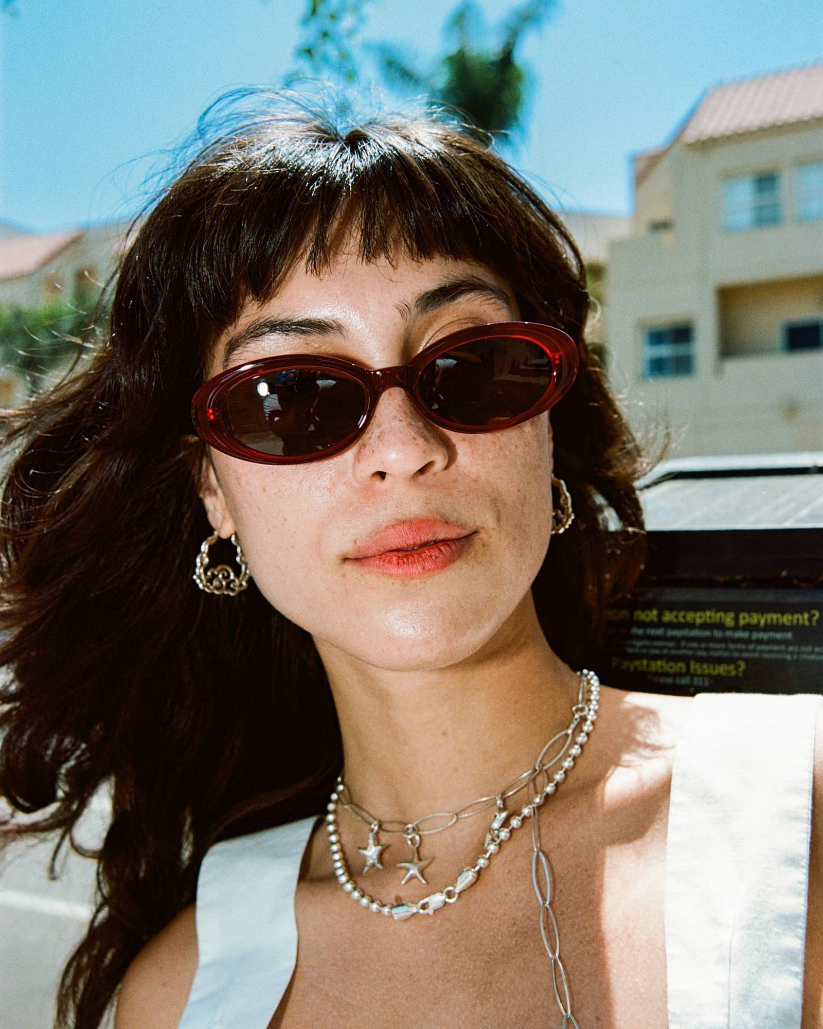 Red oval sunglasses with dark tinted lenses and tortoiseshell-style translucent frames, worn outdoors in bright sunlight