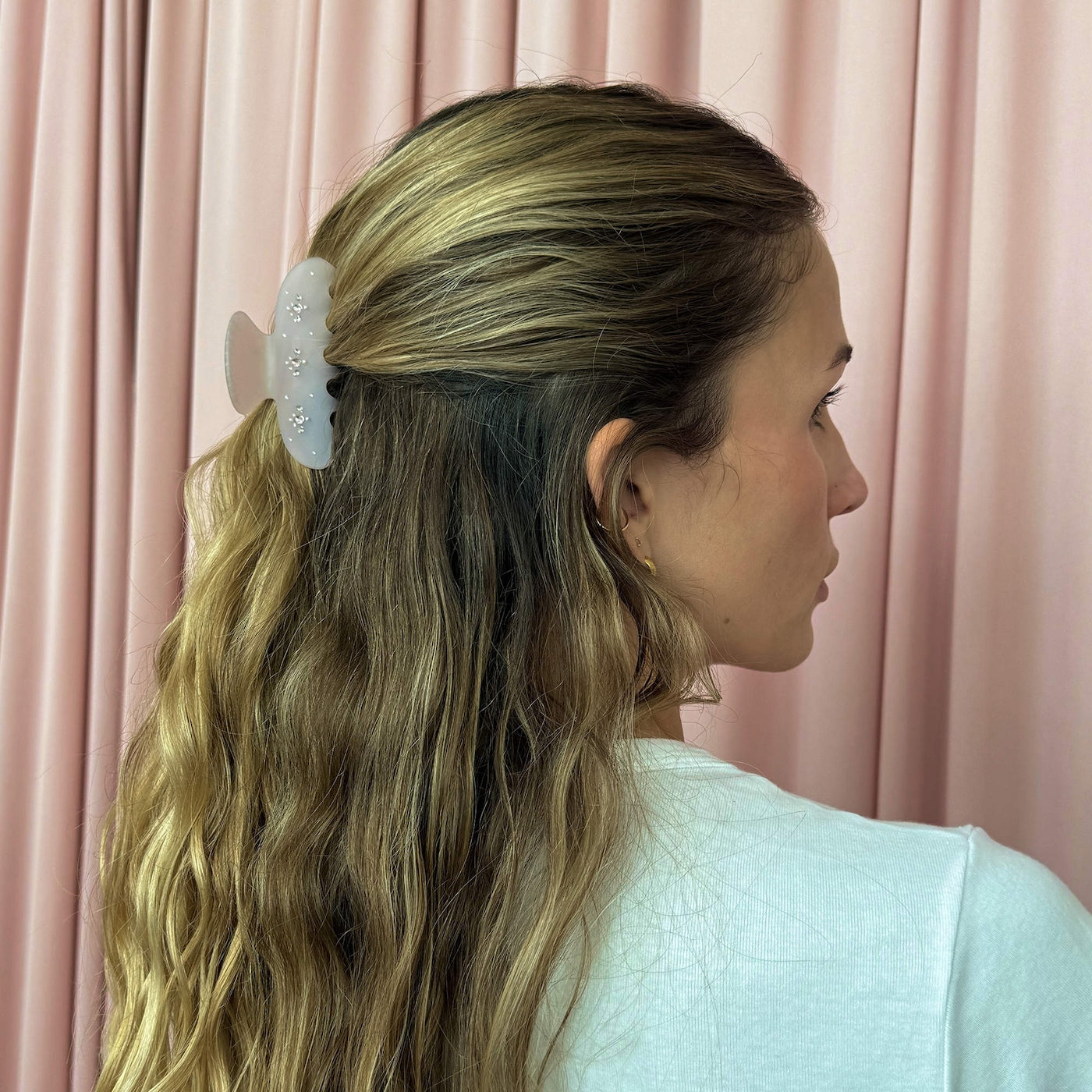 White floral hair claw clip with pearl embellishments holding blonde hair in a half-up style against pink curtain background