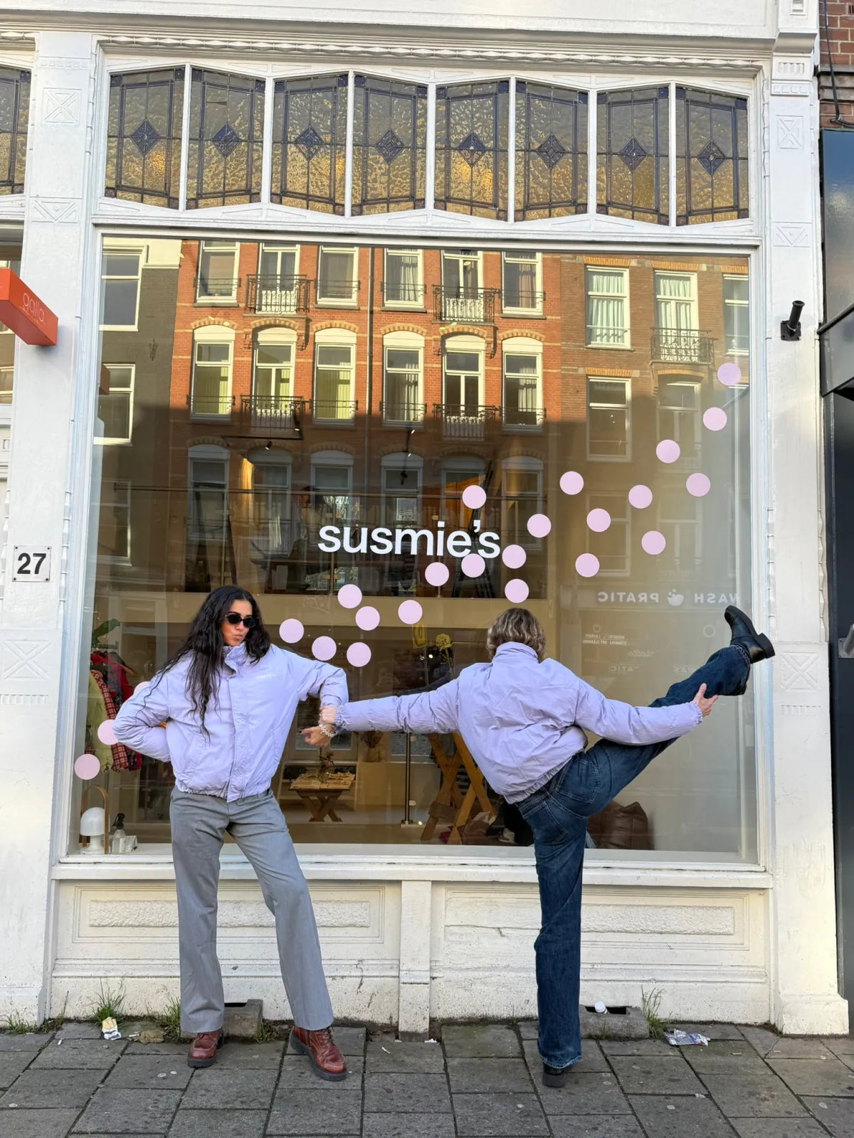 Two people in matching lavender jackets and jeans pose playfully outside a storefront with decorative stained glass windows