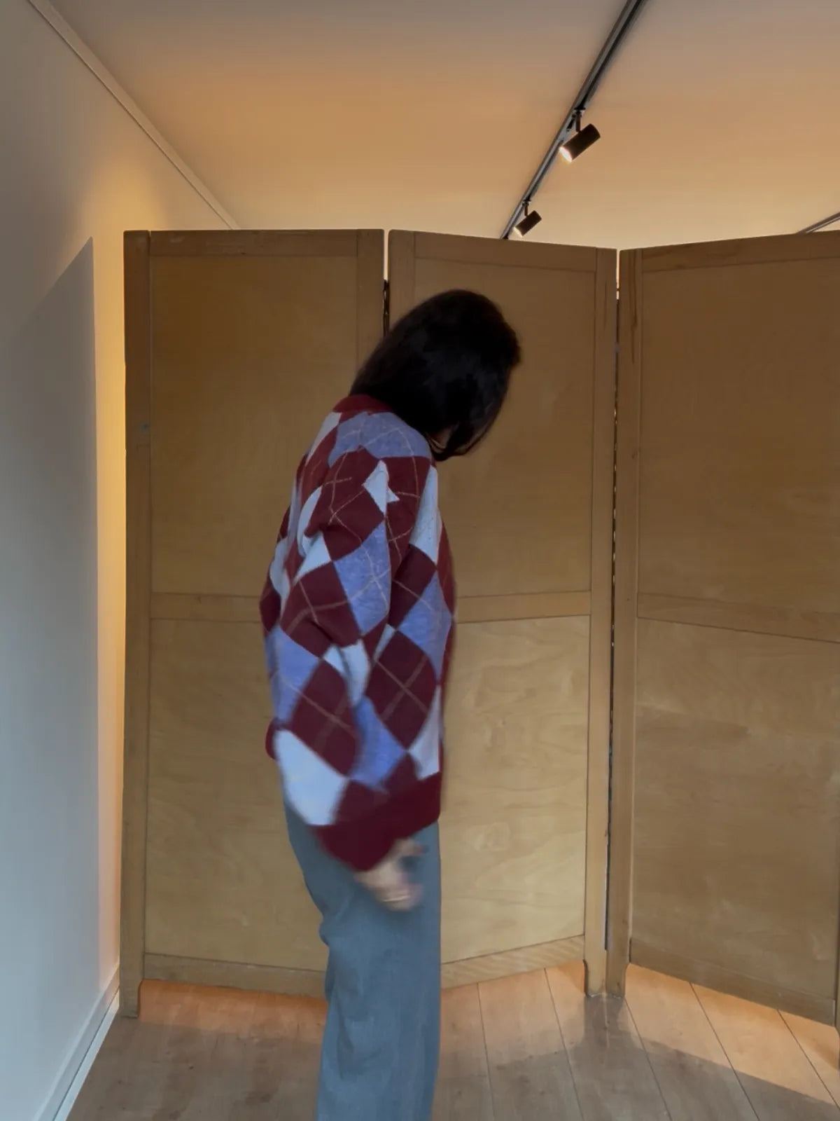 Person wearing red and white argyle knit sweater with light blue jeans in a minimalist room with wood panels