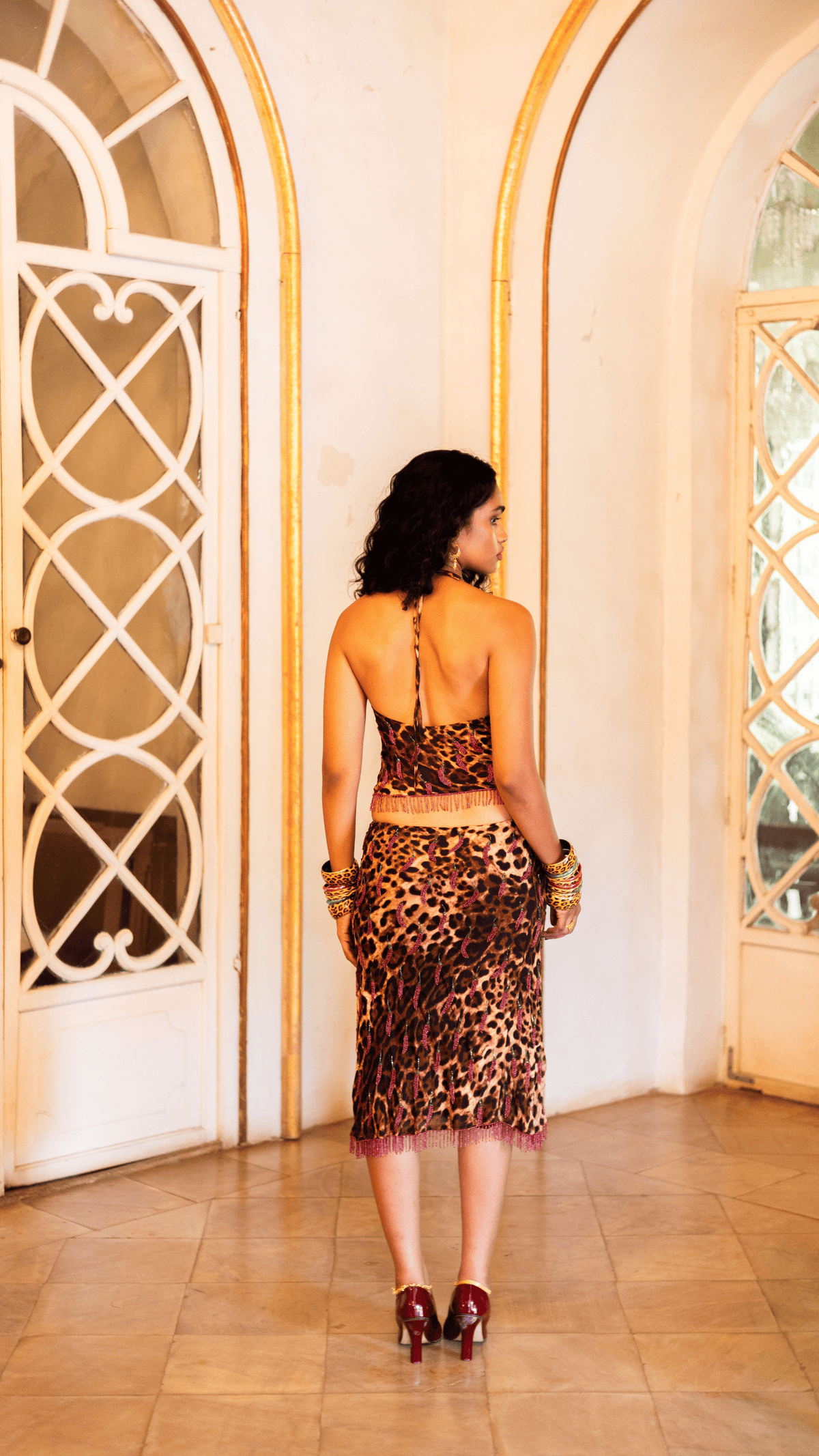 Woman in leopard print halter top and midi skirt set with fringe detail, gold bangles, and burgundy heels viewed from back