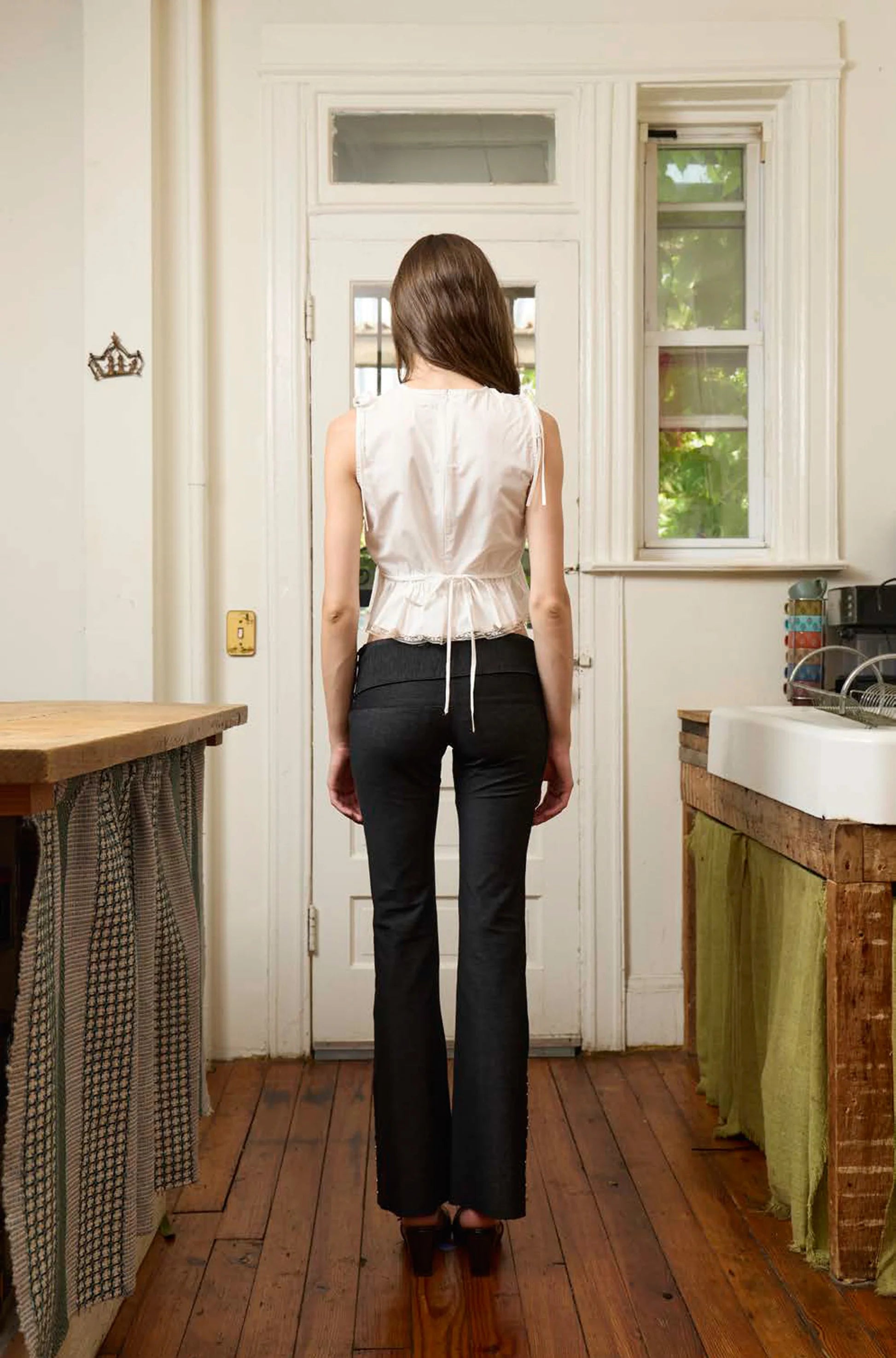 Woman wearing white sleeveless peplum top with drawstring waist and black flared pants, standing in kitchen facing door
