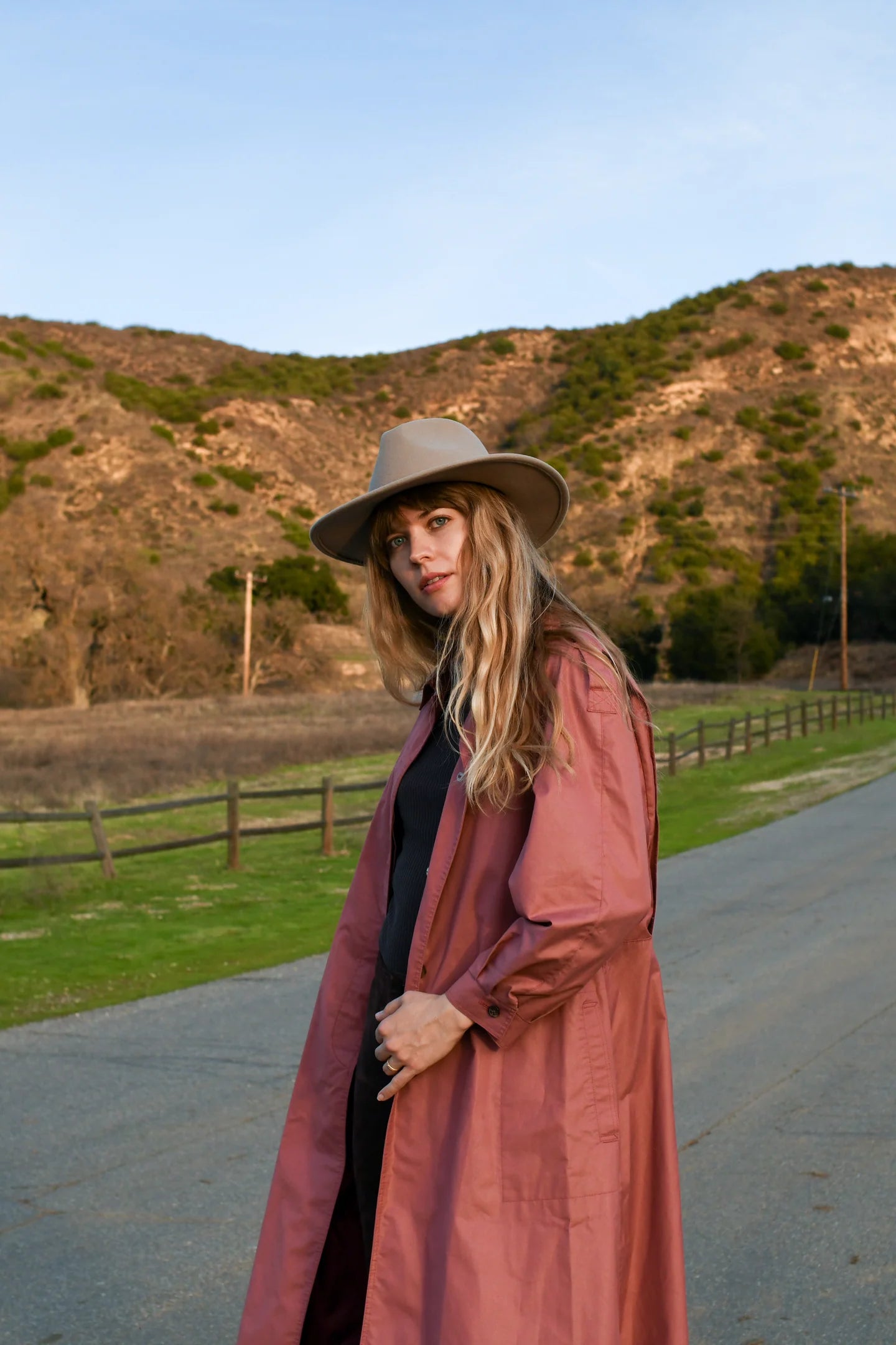 Woman wearing a long dusty rose trench coat with gray fedora hat standing on rural road with hills in background