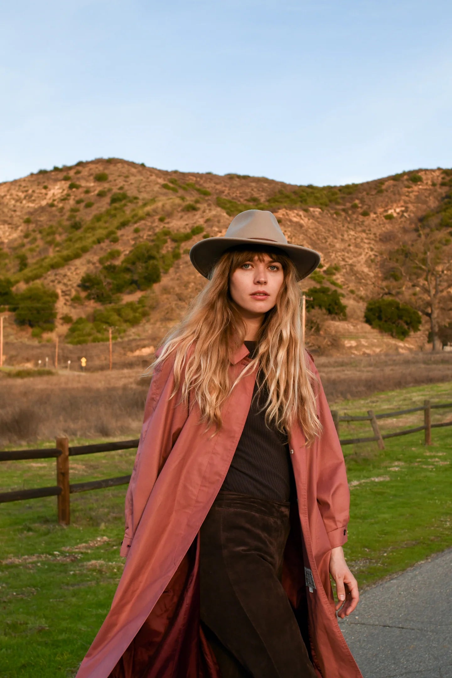 Woman wearing long rust-colored duster coat with wide-brim tan hat in outdoor setting with hills and fence in background