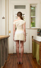 Woman wearing cream-colored short-sleeve top and pleated mini skirt with lace waistband detail, viewed from behind in rustic kitchen