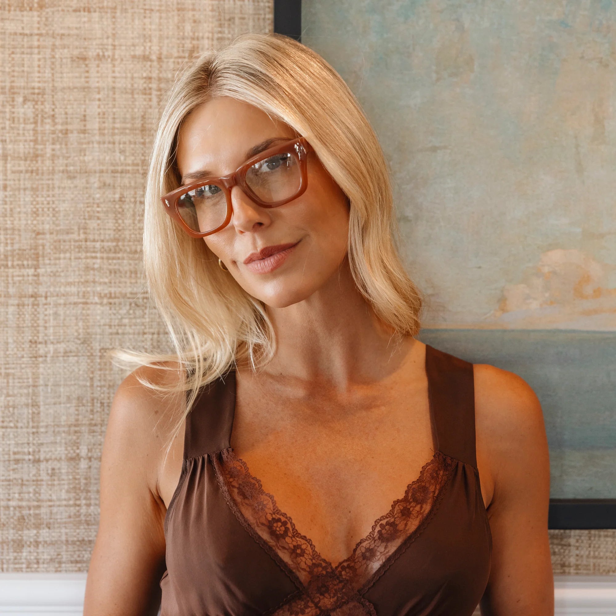 Woman wearing glasses and a brown top with lace details, sitting indoors.