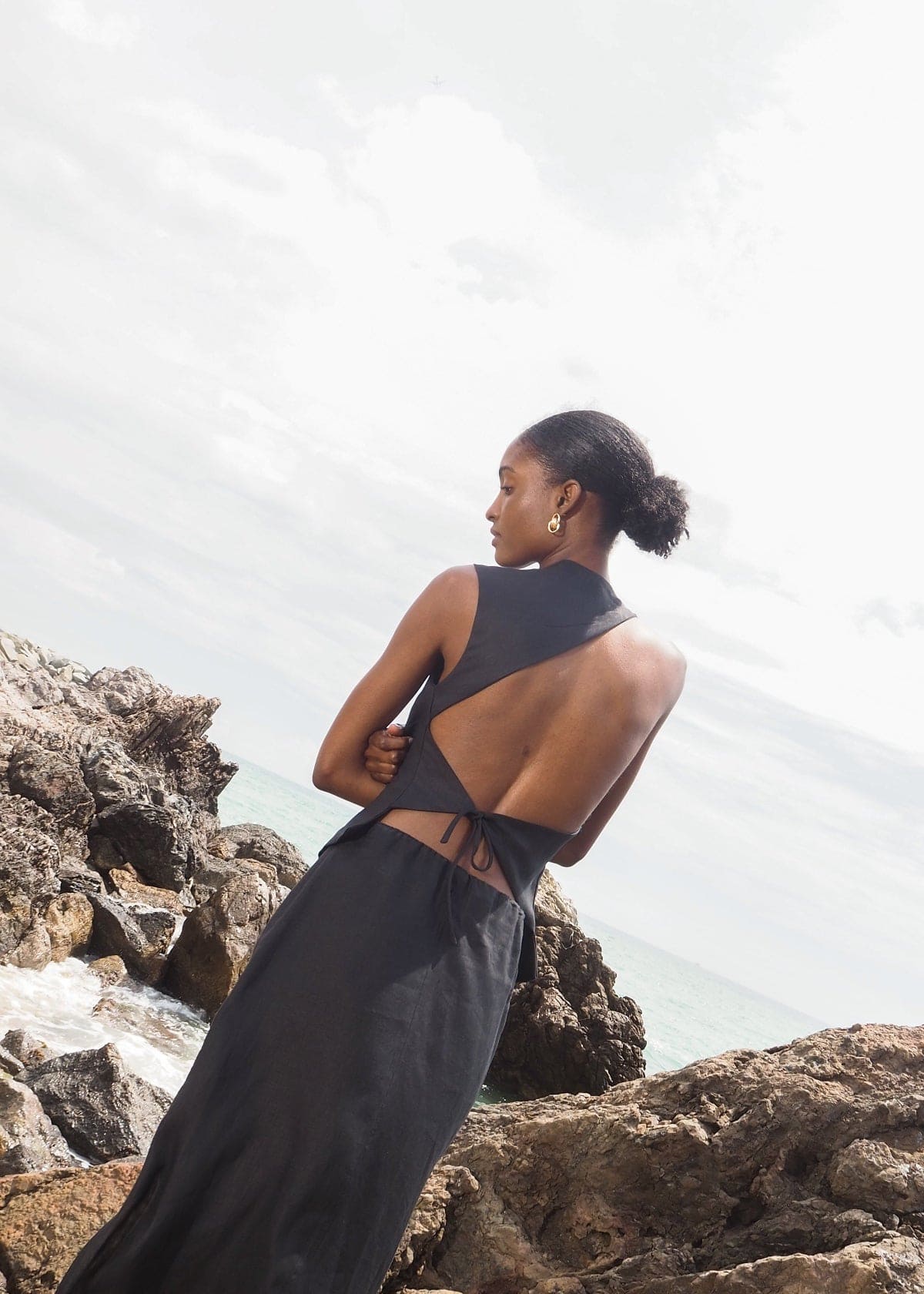 Black sleeveless maxi dress with open back cutout detail, worn by model standing on rocky coastline