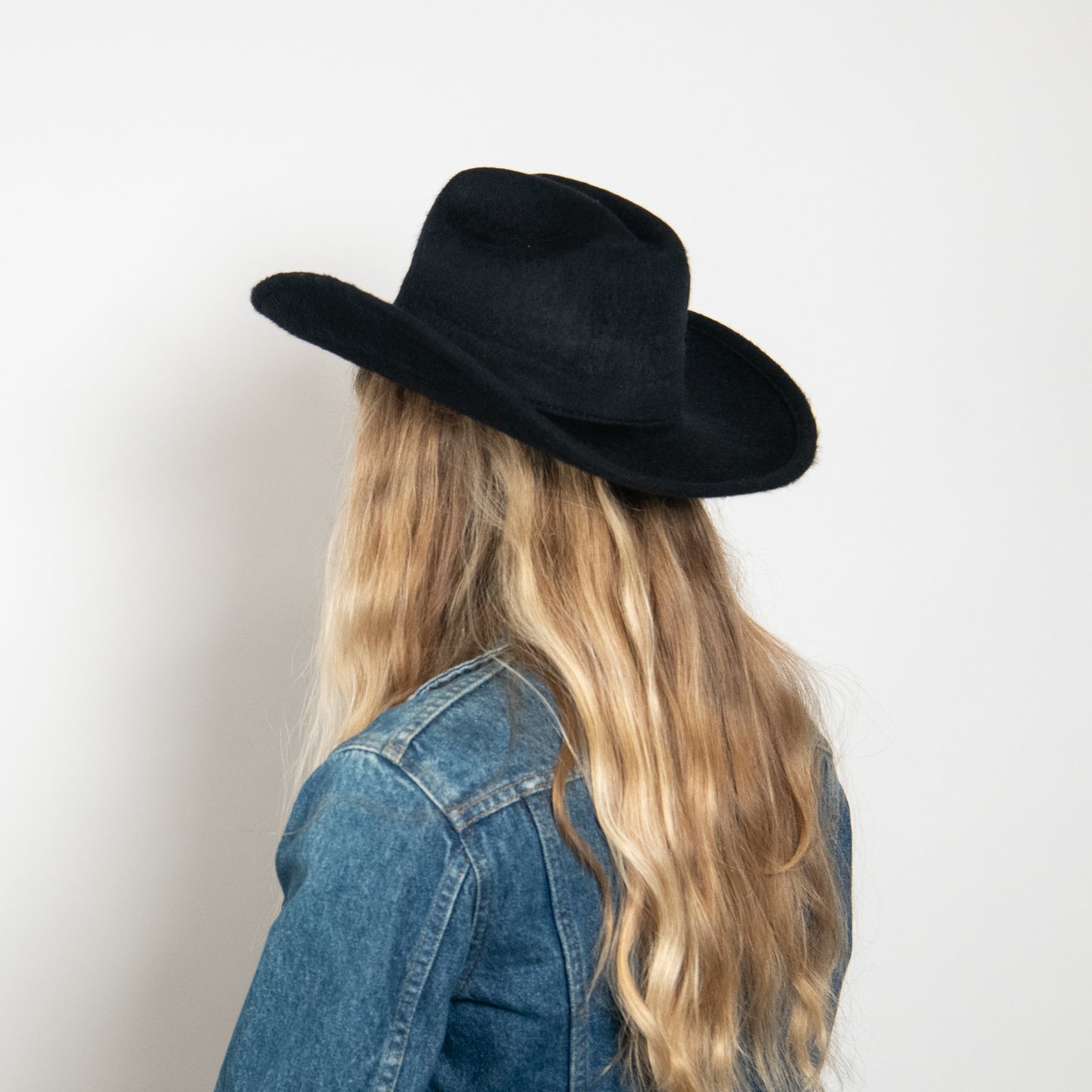 Black felt wide-brim cowboy hat worn by person with long blonde hair and denim jacket, viewed from behind