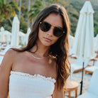Woman wearing sunglasses and a white strapless top in a tropical setting with white umbrellas and greenery.