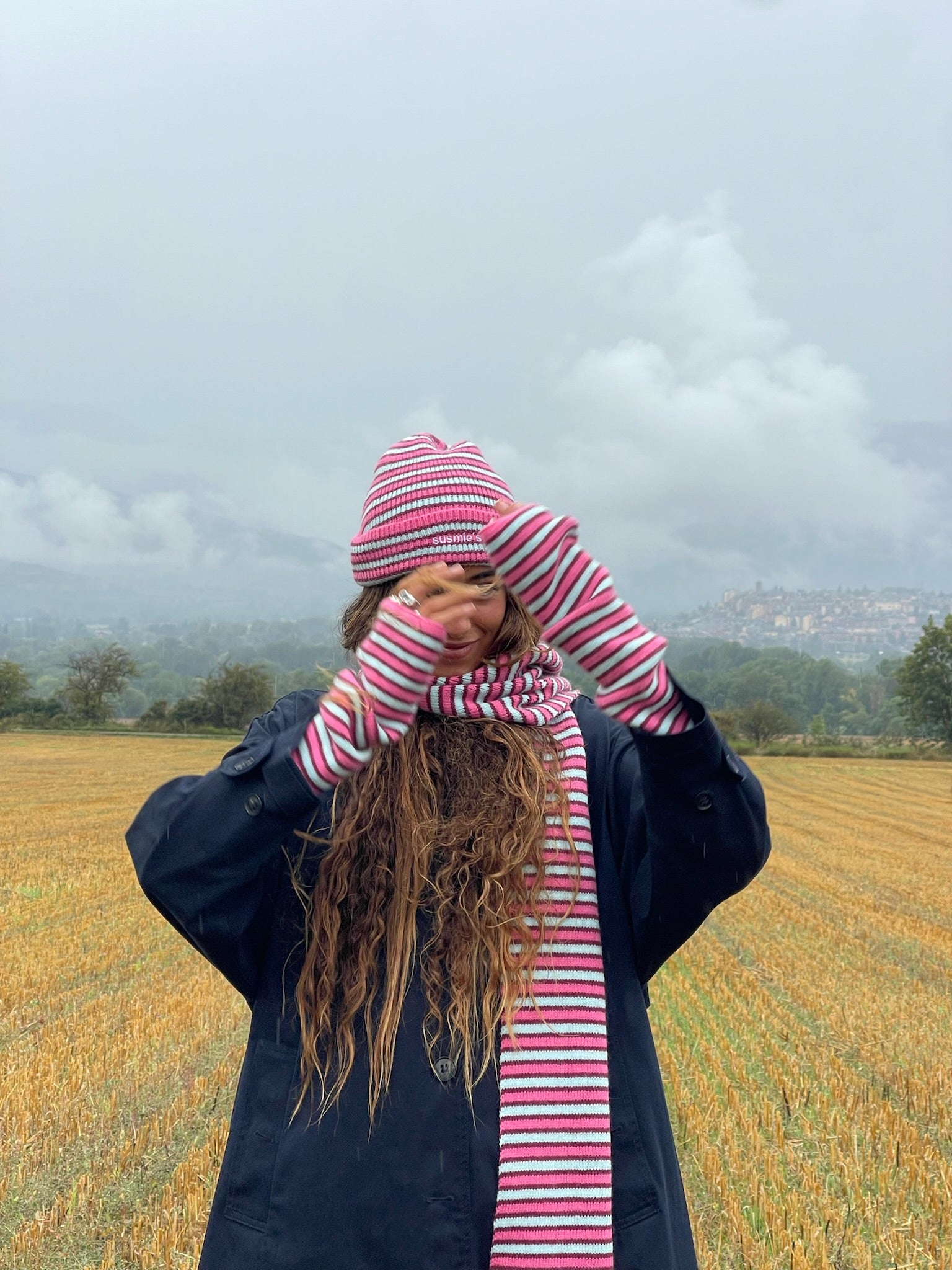 Person wearing pink and white striped knit beanie and matching scarf set in an outdoor field setting
