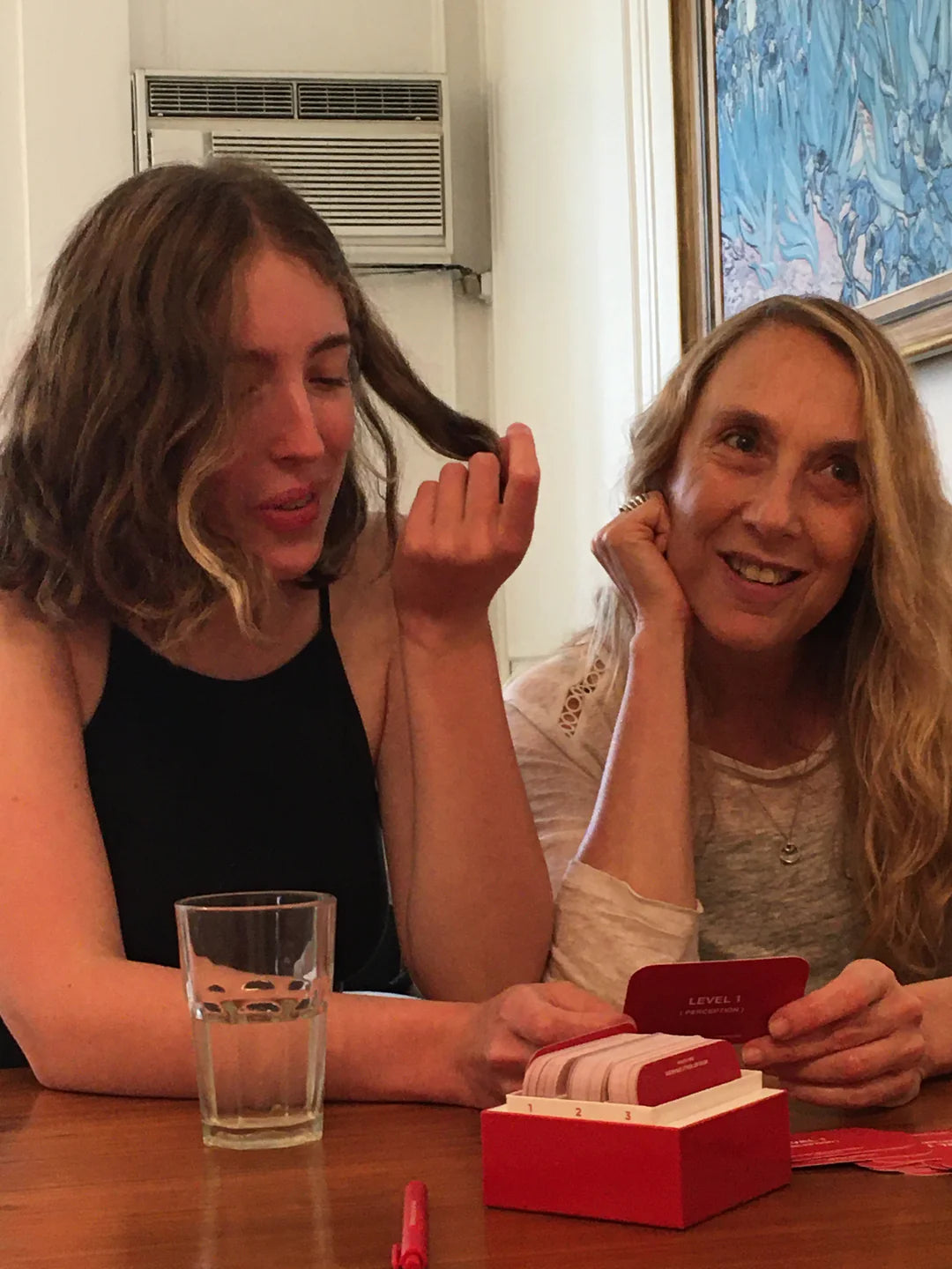 Two women playing a card game at wooden table with red game box and glass of water