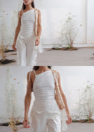 White asymmetric ruched cami top with thin straps, worn with matching white wide-leg trousers and a tan bag.