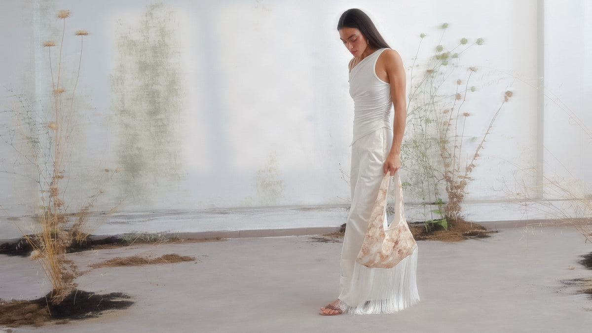 Floral printed hobo bag with fringe trim in soft beige tones, held by model in white ensemble.