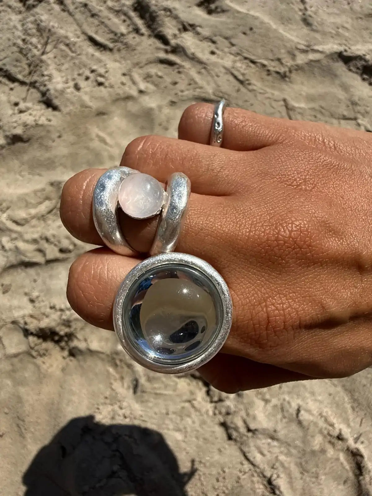 Hand wearing three silver rings with rose quartz and googly eye stones on sandy beach background