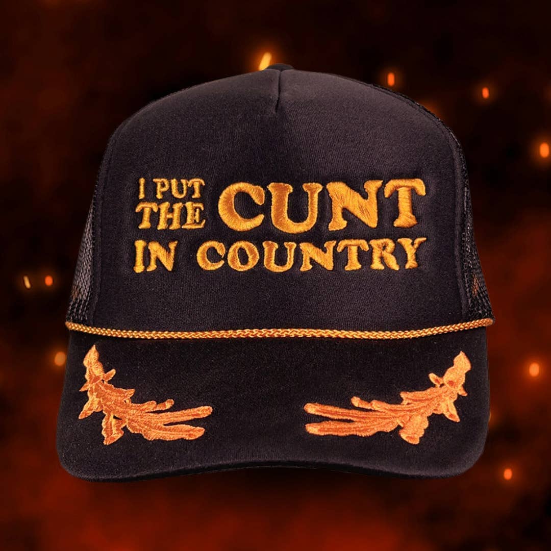 Black trucker hat with orange embroidery reading "I PUT THE CUNT IN COUNTRY" and witch decorations on the brim
