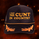 Black trucker hat with orange embroidery reading "I PUT THE CUNT IN COUNTRY" and witch decorations on the brim