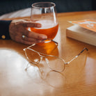 Glass of amber liquid held by a hand on a wooden table with glasses and a book.