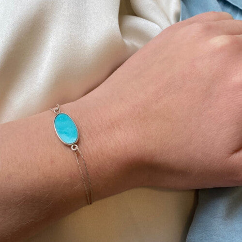 Delicate gold chain bracelet with oval turquoise stone centerpiece on wrist against white fabric background