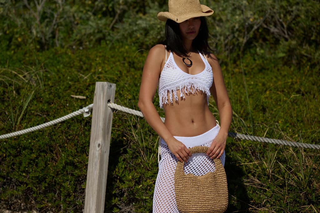 Woman in a white crochet top and skirt with a straw hat and bag, standing outdoors.