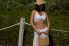 Woman in a white crochet top and skirt with a straw hat and bag, standing outdoors.