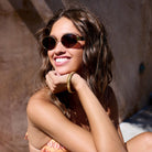 Woman wearing round gold-framed sunglasses with dark lenses and gold bangle bracelet, smiling in sunlight
