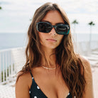 Woman wearing large rectangular black sunglasses with teal-tinted frames at the beach, long brown hair, gold necklace, polka dot bikini