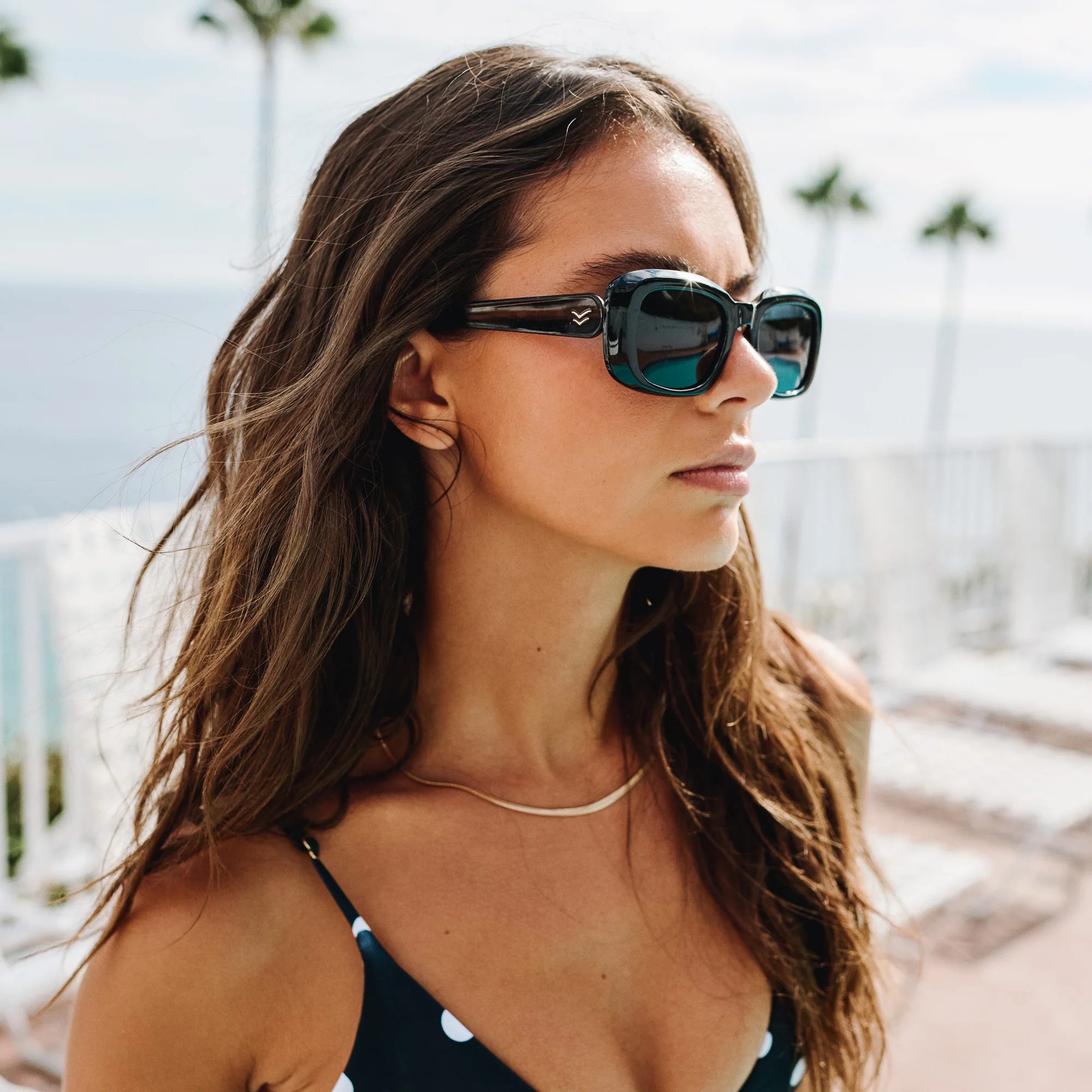 Woman wearing oversized blue square sunglasses with dark lenses and gold necklace in outdoor beach setting