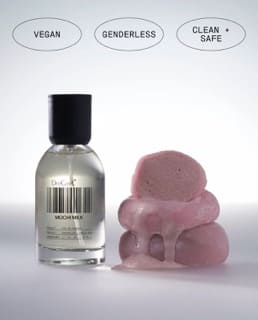 Clear glass bottle with black cap next to pink foam hair rollers, labeled vegan, genderless, and clean + safe