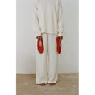 Person wearing cream oversized knit sweater and matching wide-leg pants, holding red woven mesh slippers by their sides