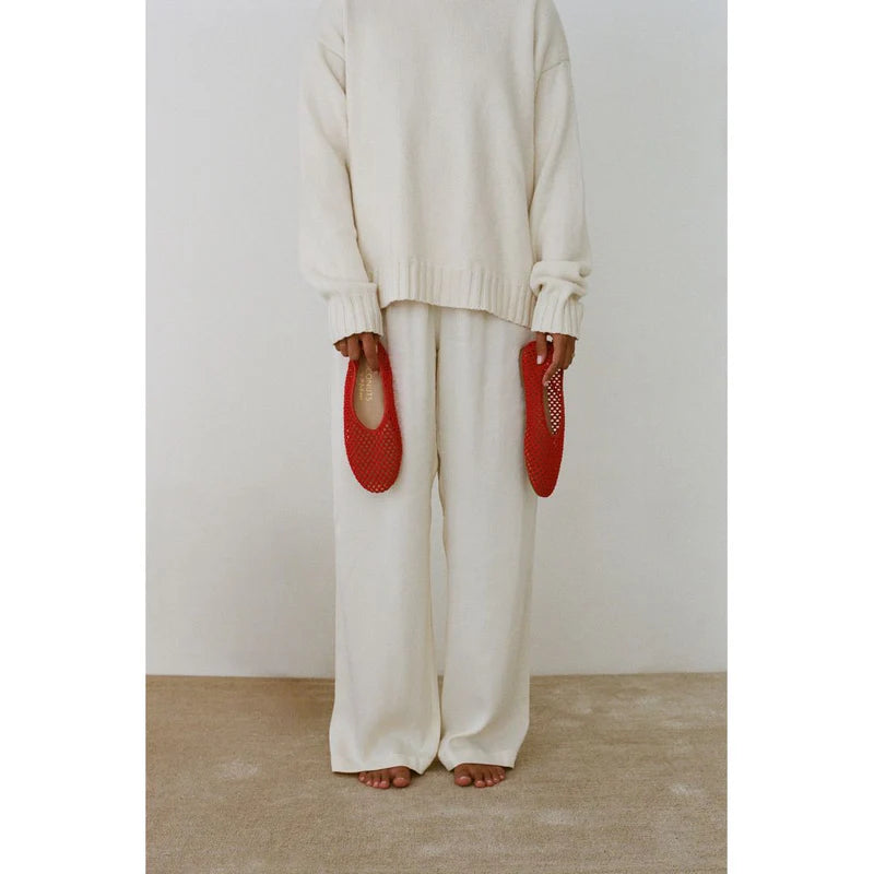Person wearing cream oversized knit sweater and matching wide-leg pants, holding red woven mesh slippers by their sides