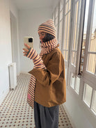 Woman wearing brown striped knit beanie, scarf, and fingerless gloves set with matching pattern, tan coat, and gray pants