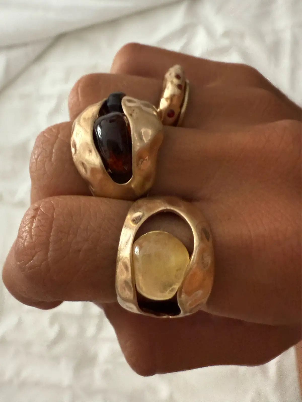 Gold statement rings with dark burgundy and pale yellow gemstones on hand against white fabric background