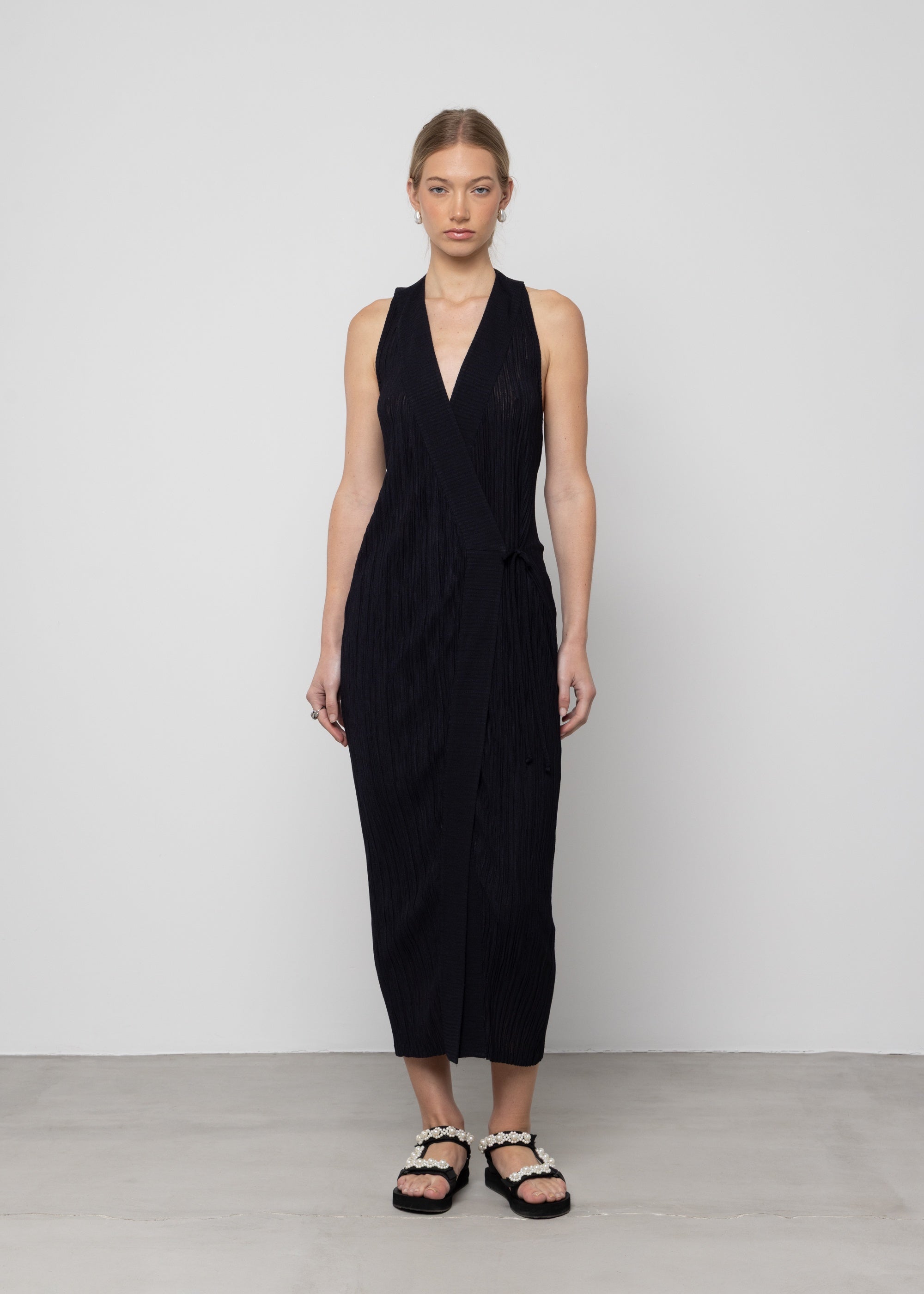 Black sleeveless midi wrap dress with textured fabric, V-neckline, and side tie detail worn with embellished sandals