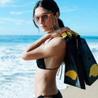 Woman wearing black bikini and orange sunglasses holding wetsuit at beach with blue ocean background