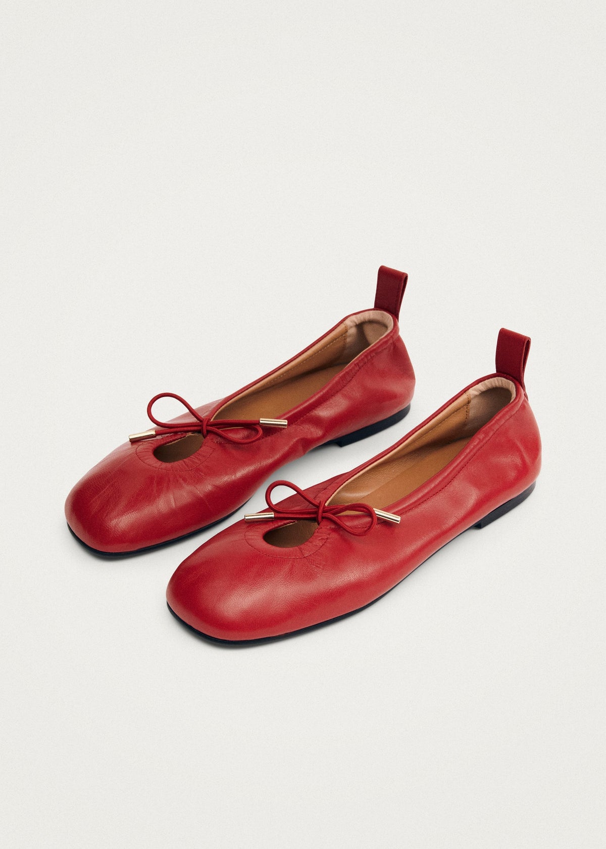 Red leather ballet flats with bow detail, gold-tipped laces, elasticated topline, and pull tab heel.