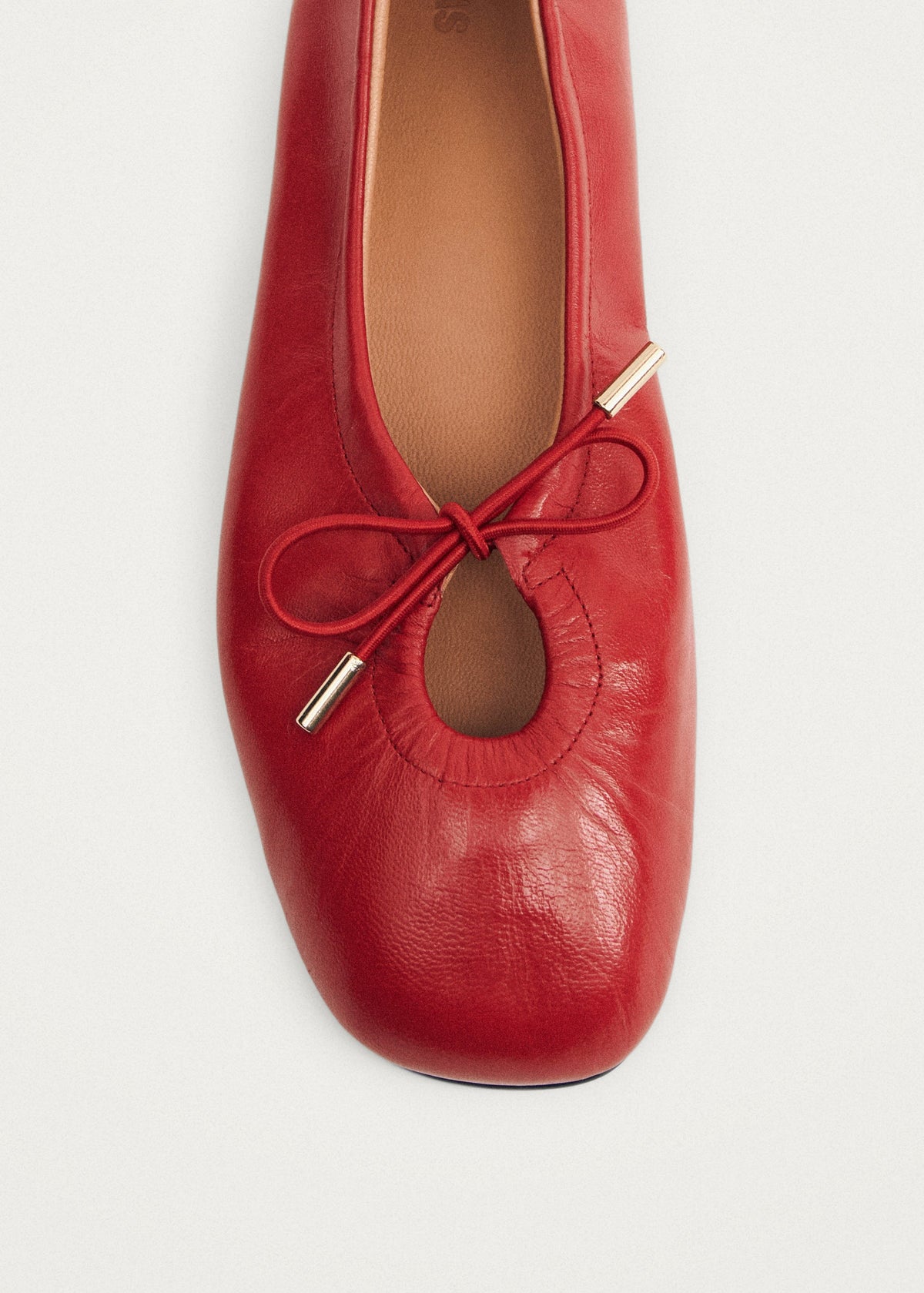 Red leather ballet flat with square toe, elastic bow detail, gold metal aglets, and tan leather insole.