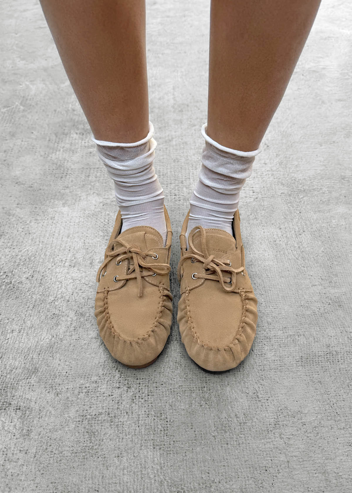 Tan suede moccasin slippers with lace-up detail, worn with white ruched ankle socks on gray surface