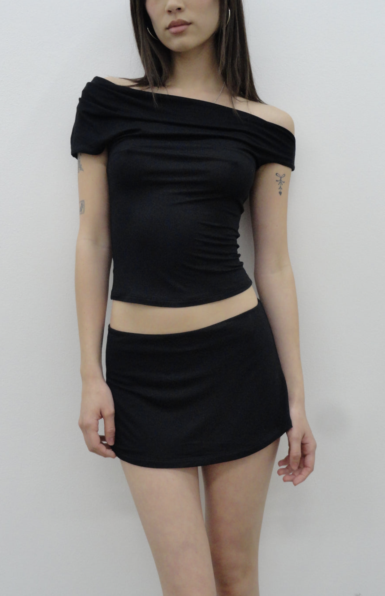 Black off-shoulder crop top and mini skirt set in stretchy fabric, worn by model with tattoos against white background
