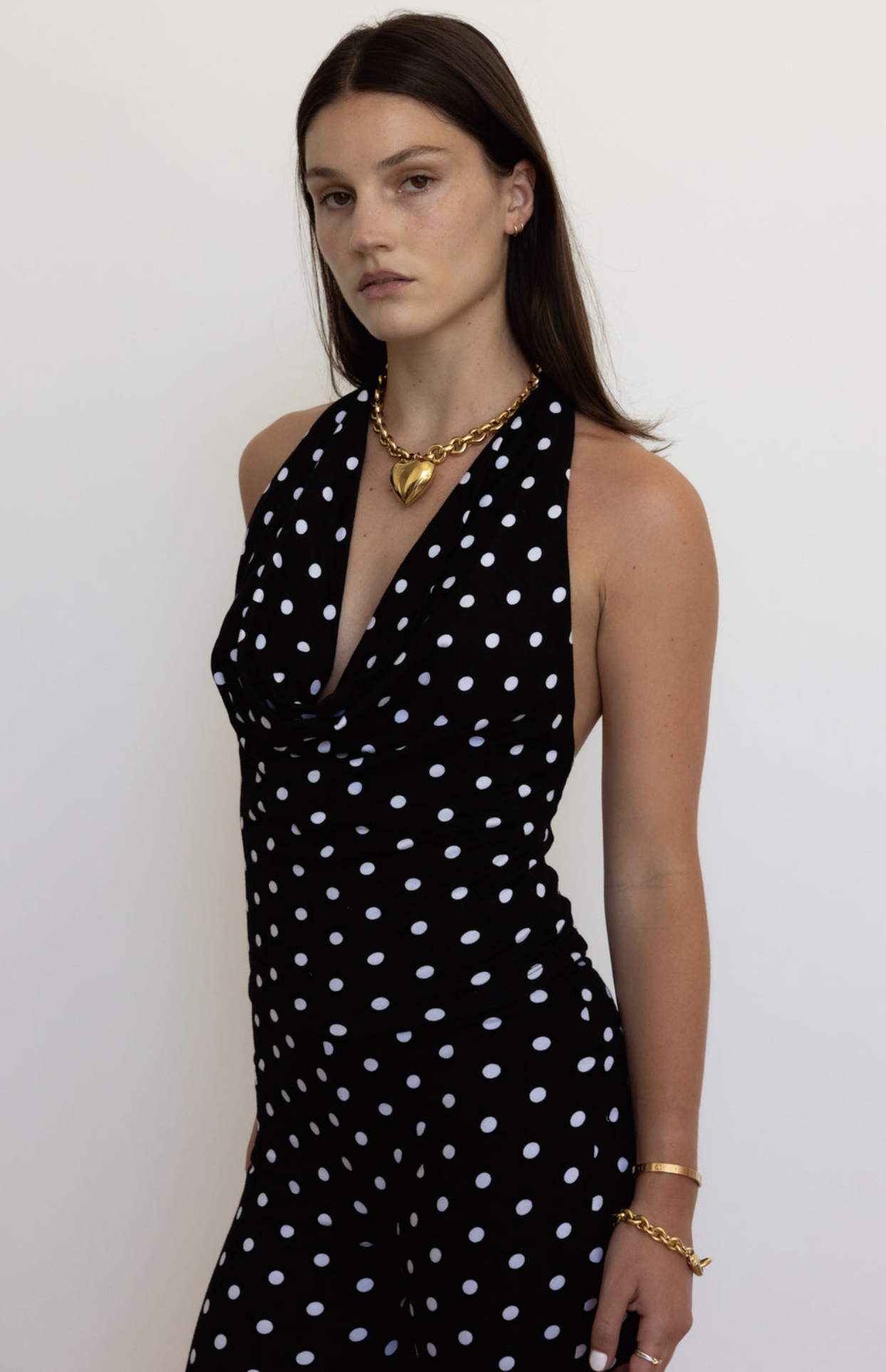 Black polka dot sleeveless dress with cowl neckline, styled with gold heart pendant necklace and chain bracelet