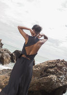Woman in black backless dress with strappy waist detail standing on coastal rocks by ocean waves