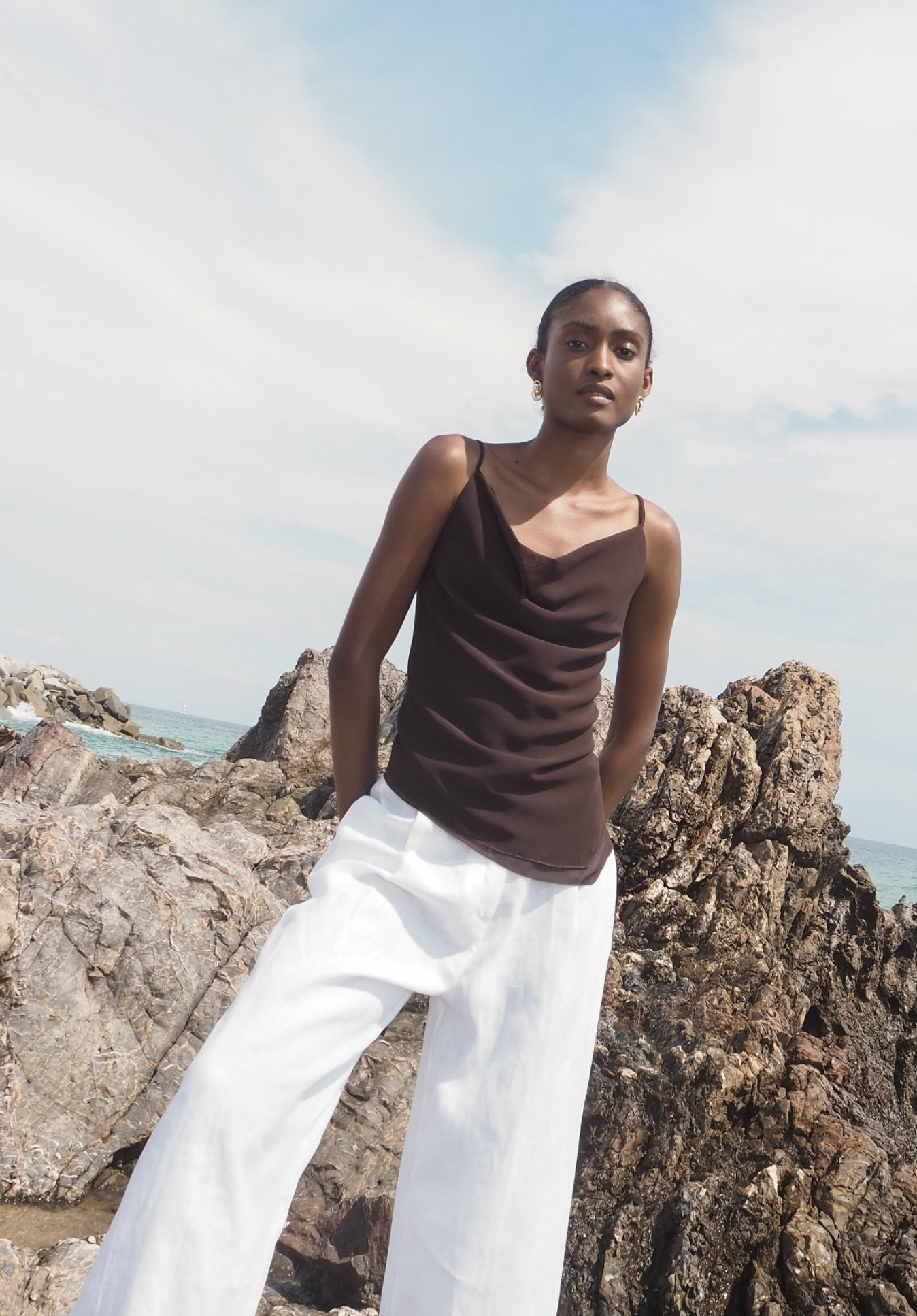 Woman wearing brown satin cowl neck camisole top and white wide-leg linen pants standing on rocky coastline