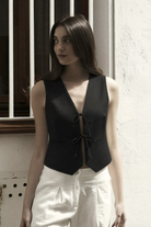 Black sleeveless vest with V-neck and tie-front closure, paired with white wide-leg pants in minimalist style