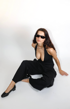 Woman in black halter bodysuit with deep V-neckline, black joggers, sunglasses, and silver choker necklace sitting on white background