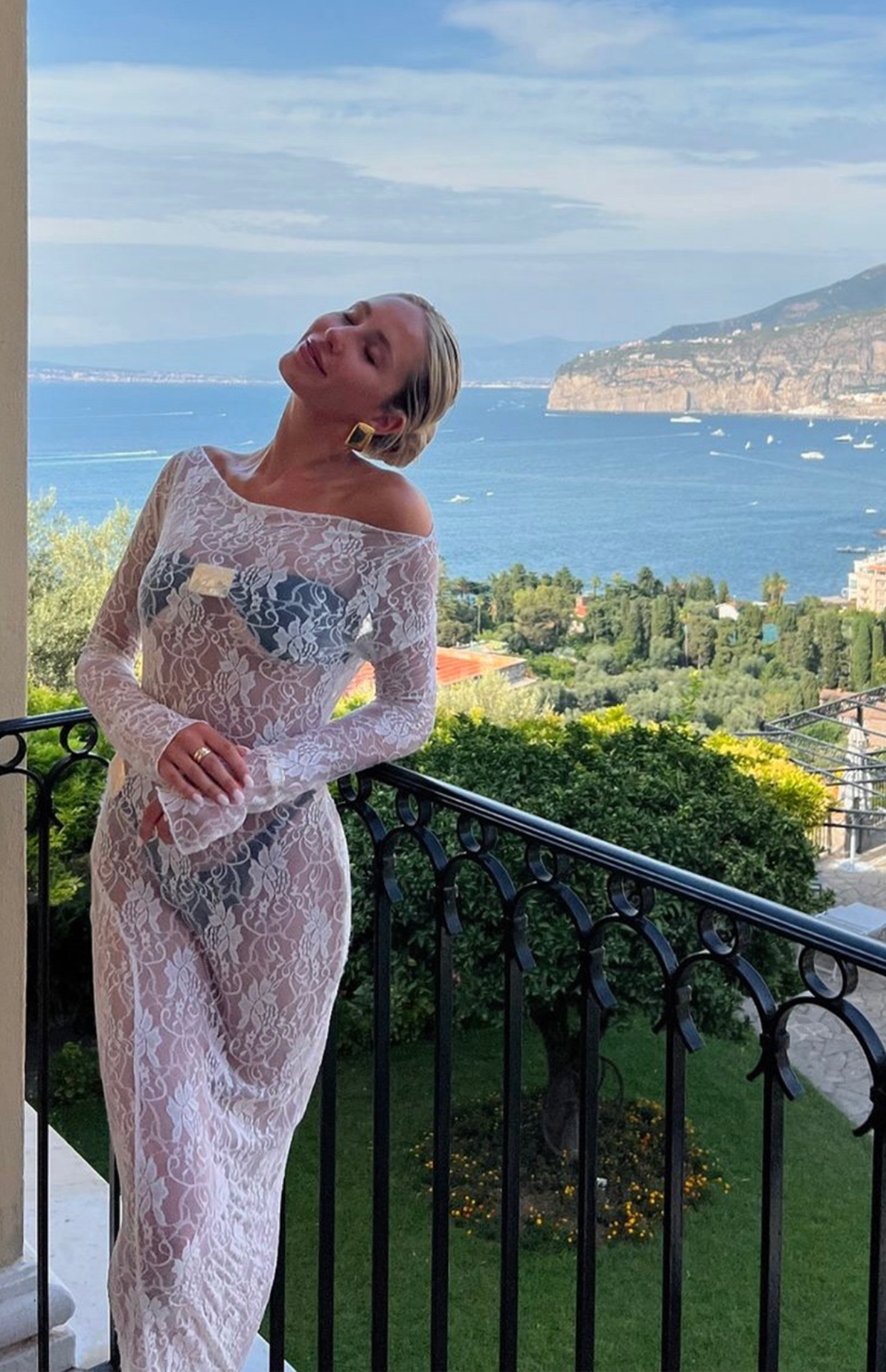 Woman in white lace off-shoulder maxi dress on balcony overlooking coastal Mediterranean scenery with blue sea and cliffs