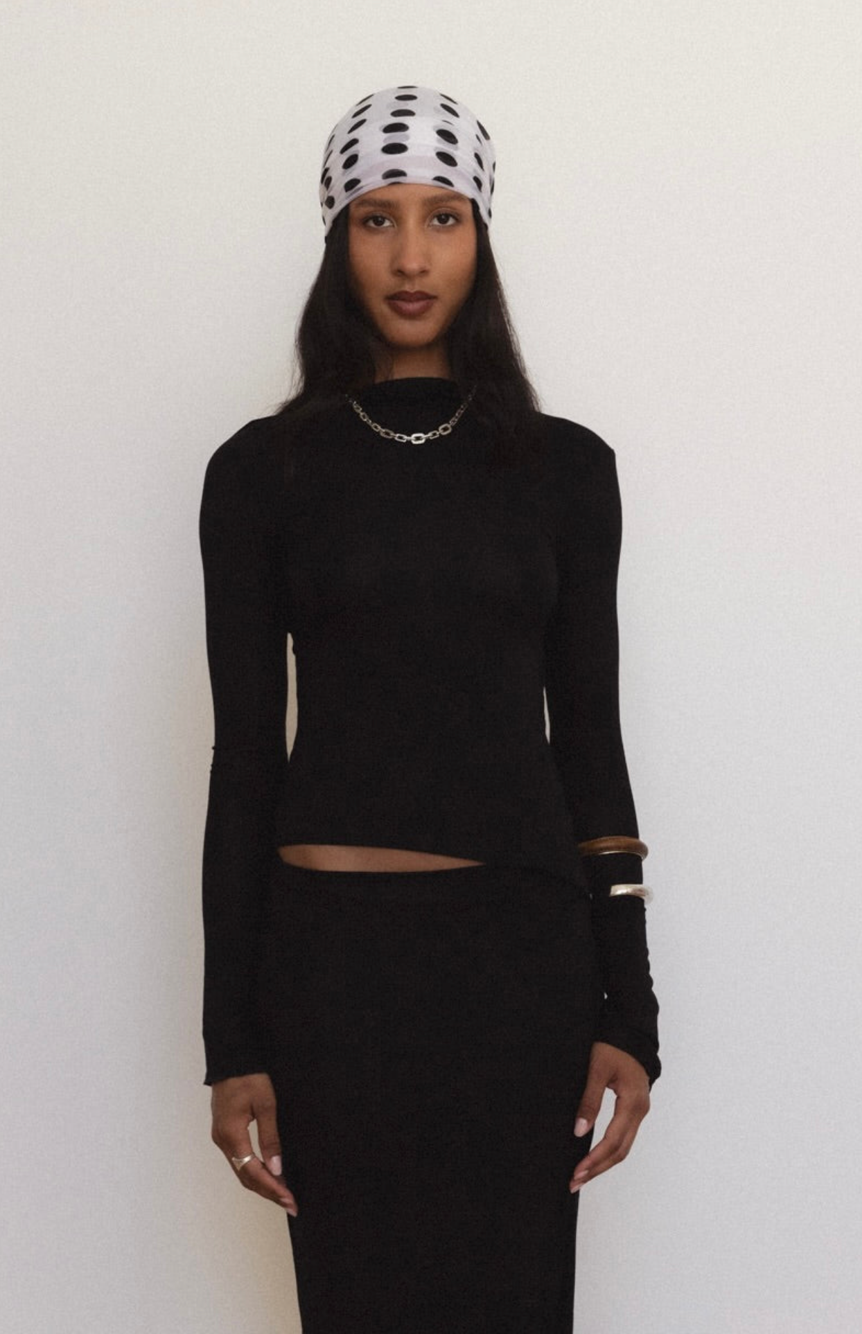 Woman wearing black long-sleeve crop top with cutout detail and fitted black skirt, accessorized with polka dot beanie and chain necklace